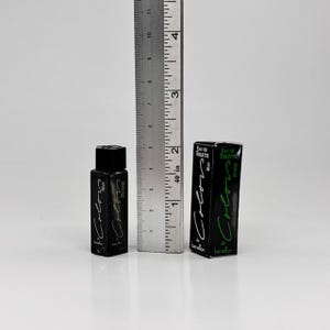 May include: A black perfume bottle and box with the text "Colors Man Homme" in green and white. The bottle is approximately 2 inches tall, and the box is about 3 inches tall. A metal ruler is next to the perfume.