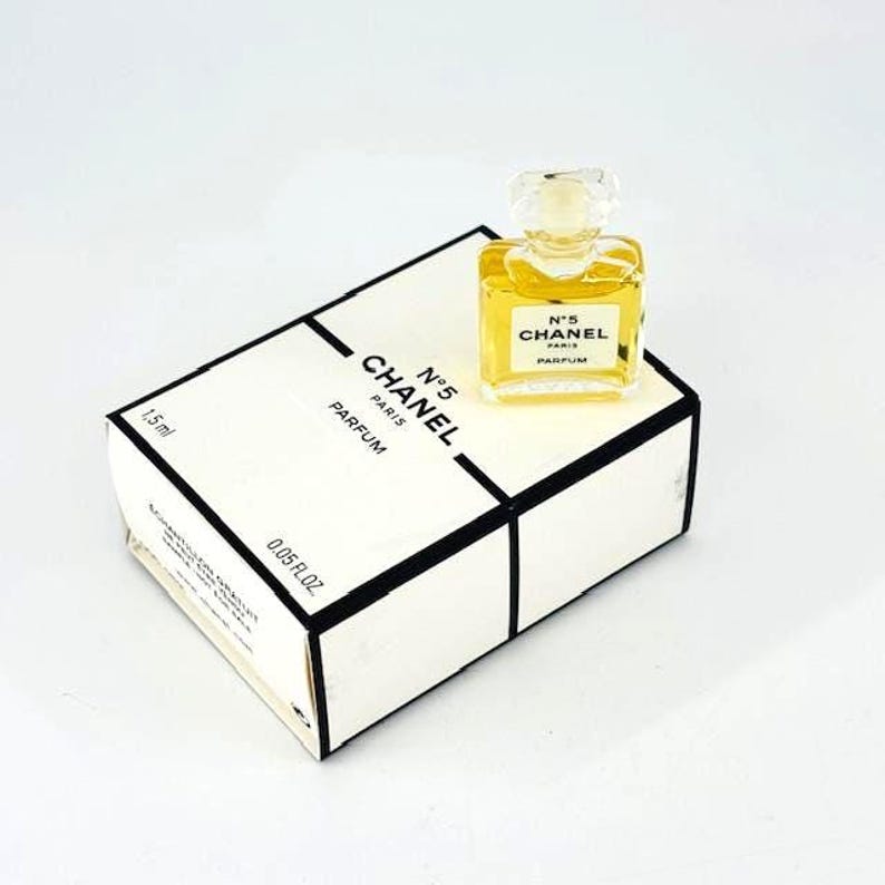 Op de afbeelding: Een kleine fles Chanel No. 5 parfum, met een doorzichtige stopper, staat op de wit-zwarte doos. De doos is gemarkeerd met "CHANEL N&deg;5 PARFUM" en bevat 1,5 ml parfum. De parfumfles is vierkant en bevat een gouden vloeistof.