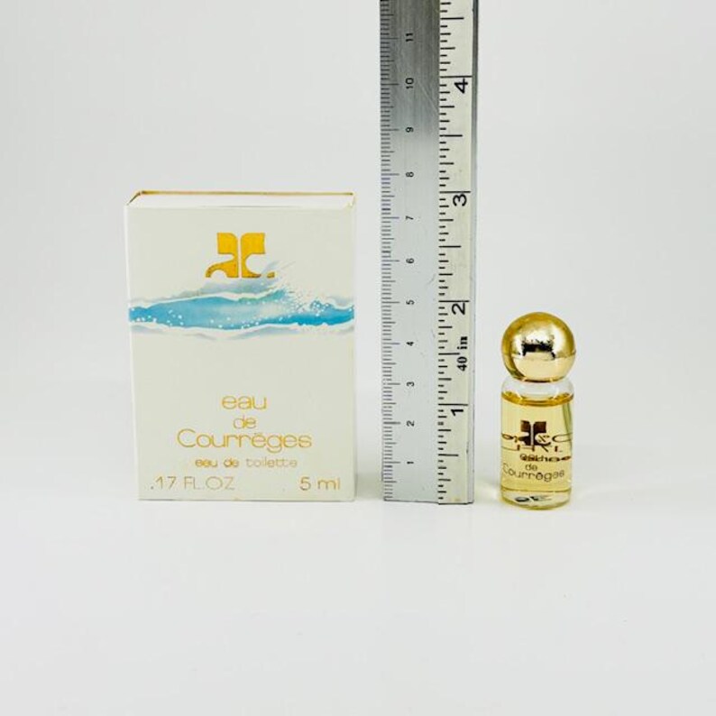 Puede incluir: Un juego de botella y caja de perfume en miniatura. La caja es blanca con gr&aacute;ficos de olas azules y texto dorado. El frasco de perfume es peque&ntilde;o, con una tapa dorada y un cuerpo de vidrio transparente. Una regla est&aacute; al lado del perfume.