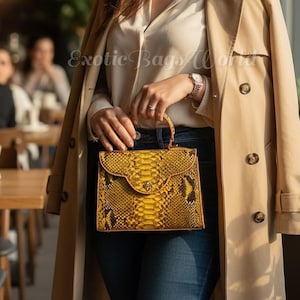 May include: A yellow and brown snakeskin handbag with a top handle is held by a person wearing a beige trench coat and blue jeans. The bag has a flap closure and a gold clasp. The background is a cafe setting.