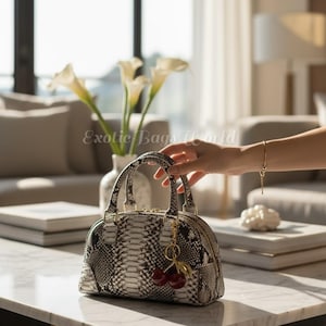 May include: A dome-shaped handbag with a snakeskin pattern in shades of gray and white. The bag has two handles and a gold zipper. A cherry charm hangs from the handle. The text "Exotic Bags World" is visible in the background.