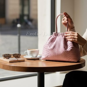 以下が含まれることがあります： ピンク色のヘビ柄ハンドバッグ。上部にハンドルが付いています。バッグは丸い木製のテーブルの上にあり、コーヒーカップとサングラスが隣にあります。「Exotic Bags」の文字が見えます。