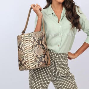 Pode incluir: Bolsa cilíndrica com padrão de pele de cobra em tons de castanho, bege e creme. A bolsa tem uma alça de couro bege e acabamento a condizer. A bolsa é segurada por uma pessoa que usa uma camisa verde clara com botões e calças estampadas.