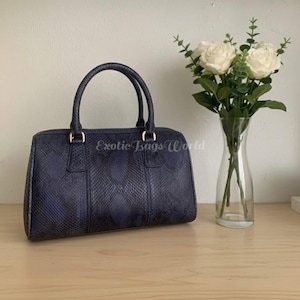 May include: A navy blue handbag with a snakeskin pattern, featuring two handles and gold-colored hardware. The bag has a rectangular shape and a zipper closure. The text "Exotic Bags World" is printed on the bag. A vase of white roses sits next to the bag.
