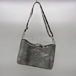 Può includere: Borsa a tracolla argentata con motivo a pelle di serpente. La borsa ha una forma rettangolare con la parte superiore curva e una lunga tracolla regolabile. La tracolla e la borsa sono attaccate con elementi metallici argentati. Il testo "exotic Bags World" è visibile.