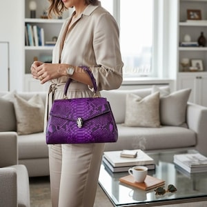 Könnte beinhalten: Eine leuchtend lila Handtasche mit Schlangenmuster. Die Tasche hat einen oberen Griff, einen silbernen Verschluss und goldfarbene Hardware. Die Tasche wird von einer Person in einem beigen Outfit gehalten.