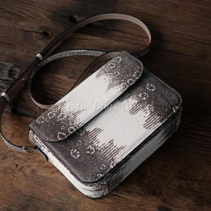 Könnte beinhalten: Rechteckige Umhängetasche mit Klappverschluss. Die Tasche hat ein weißes und graues Muster mit passendem Riemen. Die Tasche besteht aus einem strukturierten Material und liegt auf einer Holzoberfläche.