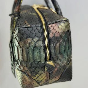 May include: A rectangular handbag with a camouflage pattern in shades of green, brown, and gold. The bag features a gold zip and handles. The texture of the bag appears to be snakeskin.