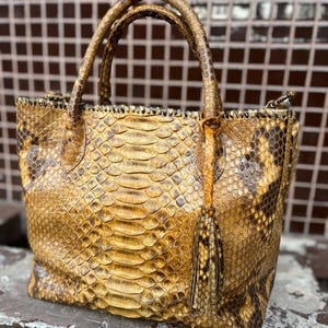 May include: A handcrafted tote bag made from genuine snakeskin. The bag features a structured design with two top handles and a decorative tassel. The snakeskin has a natural pattern in shades of brown and gold.