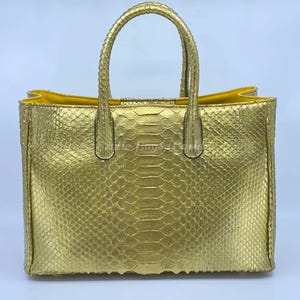 Peut inclure: Un sac fourre-tout doré avec un motif peau de serpent. Le sac a deux poignées et un intérieur jaune. Le sac est rectangulaire avec un design structuré, adapté au transport de divers articles.