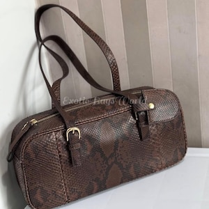 Könnte beinhalten: Eine braune Handtasche aus Schlangenleder mit rechteckiger Form und zwei Henkeln. Die Tasche hat goldfarbene Reißverschlüsse und Schnallen. Die Tasche ist aus einem Schlangenmuster mit Braun- und Schwarztönen gefertigt.