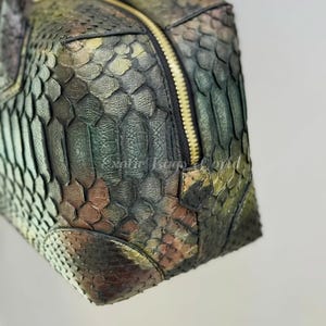 May include: A close-up of a textured, multi-coloured handbag. The bag features a zip closure and a braided handle. The colours include shades of green, brown, and gold, creating a unique, eye-catching design.