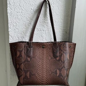 May include: A brown tote bag with a snakeskin pattern. The bag has two long handles and a button closure. The bag is made of a textured material and has a rectangular shape. The bag is hanging against a white wall.