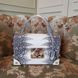 Könnte beinhalten: Eine Handtasche mit weißem und grauem Muster mit einem oberen Griff und einem goldfarbenen Verschluss. Die Tasche hat eine rechteckige Form mit abgerundeten Ecken und goldenen Nieten. Das Muster ähnelt einem Schlangenhaut-Design.