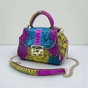 May include: A small, colorful handbag with a snakeskin pattern. The bag features a top handle and a gold chain strap. The color scheme includes purple, blue, yellow, and pink. The bag has a gold clasp closure.