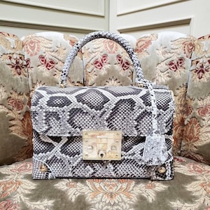 Può includere: Una borsa con motivo a pelle di serpente con manico superiore e chiusura dorata. La borsa presenta una chiusura con patta e un nappa decorativa. La borsa è appoggiata su una sedia con motivo floreale.