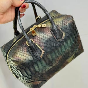 May include: A multi-coloured handbag with a snakeskin pattern. The bag has a rectangular shape, a top zip, and two handles. Gold-coloured hardware accents the bag, including the zip and handle attachments. The bag's colours include green, brown, and gold.