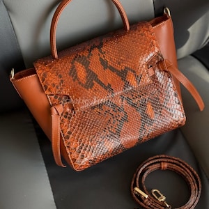 Peut inclure: Sac à main marron et orange à motif peau de serpent, avec une poignée supérieure et une bandoulière amovible. Le sac est doté d'une fermeture à rabat et est en cuir. Le sac est de style sacoche.