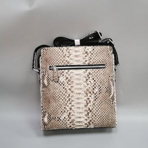 May include: A rectangular crossbody bag with a snakeskin pattern in shades of brown and white. The bag has a black zip, a black adjustable strap, and a white accent. The bag is designed to be worn over the shoulder.