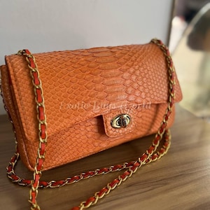 Op de afbeelding: Oranje handtas met een slangenleertextuur. Het heeft een gouden sluiting en een kettingriem met rode lederen details. De tekst "Exotic Bags World" is zichtbaar. De tas is rechthoekig van vorm.