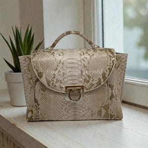 May include: A beige handbag with a snakeskin pattern. The bag has a top handle and a flap closure with a gold-colored ring detail. The words "Exotic Bags World" are visible on the bag.