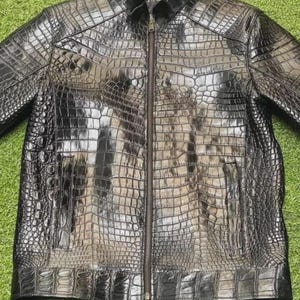 Puede incluir: Chaqueta de cuero negro con un patrón de piel de cocodrilo. La chaqueta tiene cierre de cremallera, cuello y dos bolsillos delanteros. Cuelga de una percha blanca sobre un fondo verde.