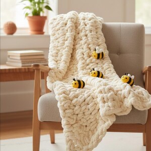 May include: A cream-colored, chunky knit blanket draped over a beige armchair. The blanket is adorned with four small, yellow and black bee-shaped decorations. The chair has wooden arms and legs, and the background includes a window and a potted plant.