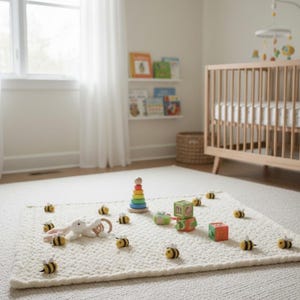 May include: A cream-colored, textured rug with a collection of baby toys. The toys include a wooden stacking tower, a plush bunny, and several small bee-shaped decorations. A wooden crib and a mobile are in the background.