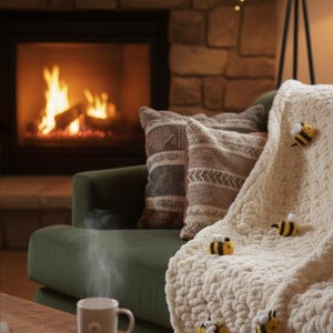 May include: A cozy living room scene with a green sofa, pillows, and a cream-colored blanket adorned with bee appliqu&eacute;s. A lit fireplace and a mug of steaming coffee create a warm, inviting atmosphere. The blanket is draped over the sofa.
