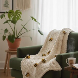 May include: A cream-colored, chunky knit blanket with several yellow and black bee appliqu&eacute;s draped over a green armchair. The blanket is soft and textured, with a cozy, handmade aesthetic. The bees are evenly spaced across the blanket.