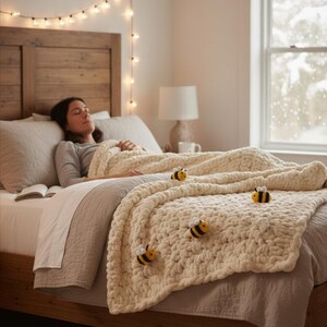 May include: A cream-colored, chunky knit blanket adorned with three bee appliqu&eacute;s rests on a bed. The bed has a wooden frame and is covered with neutral-toned bedding. A string of lights hangs above the bed, creating a cozy atmosphere.