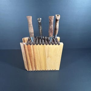 Könnte beinhalten: Ein Holz-Stifthalter mit vier handgefertigten Stiften. Der Halter besteht aus hellem Holz mit einem strukturierten, geriffelten Design. Die Stifte haben Holzschäfte in verschiedenen Brauntönen und silberne Spitzen.