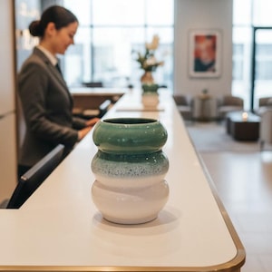 Puede incluir: Un jarrón decorativo de cerámica con diseño apilado. La sección superior es de color verde oscuro, que transiciona a una base blanca con un centro moteado verde claro. El jarrón está sobre una encimera blanca con un borde dorado, lo que sugiere un hotel u oficina.