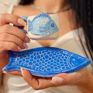 May include: A blue ceramic cup and saucer set. The cup features a blue fish design on a white background. The saucer is shaped like a fish and is blue with white detailing. The set is held in a person's hands.