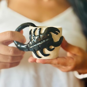 May include: A cream-coloured ceramic mug featuring a three-dimensional black scorpion sculpture attached to the exterior. The scorpion's curved tail forms the handle of the mug, while its body and legs are moulded onto the side of the vessel.