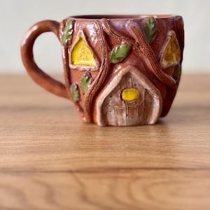 May include: A brown ceramic mug featuring a sculpted house design with a central door, a yellow circular knob, and triangular windows. The exterior is decorated with raised tree branch textures and small green leaves, finished with a glossy glaze.