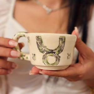 May include: Cream-colored ceramic mug with a green and black Taurus zodiac symbol. The handle and rim feature green floral accents. The mug is held in a person's hands.