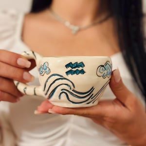 May include: Cream-colored ceramic mug with blue wave and cloud designs, and the Aquarius symbol. The mug has a curved handle and is held by a person. The mug is decorated with hand-painted blue lines and shapes.