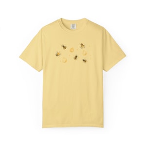 May include: A light yellow t-shirt featuring a design of bees, honeycombs, and honey droplets. The bees are black and yellow, and the honeycombs and droplets are shades of yellow and orange. The shirt is a casual, short-sleeved style.