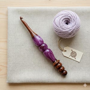 Peut inclure: Un crochet artisanal doté d'une pointe et d'une base en bois brun foncé poli, avec une section centrale en résine violette marbrée. Une pelote de laine lavande claire est accompagnée d'une petite étiquette beige avec une icône de pelote.