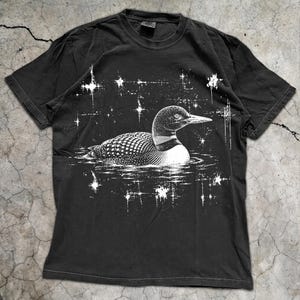 May include: A black short-sleeve t-shirt featuring a white graphic print of a loon swimming on water. The design includes a stippled texture on the bird and surrounding white starburst shapes scattered across the chest area of the garment.