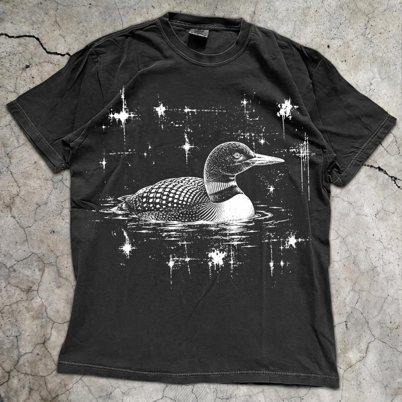 May include: A black short-sleeve t-shirt featuring a white graphic print of a loon swimming on water. The design includes a stippled texture on the bird and surrounding white starburst shapes scattered across the chest area of the garment.