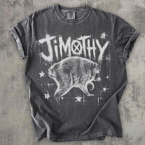 May include: A charcoal gray short-sleeve t-shirt featuring a white graphic print of a raccoon and the text "JIMOTHY" in a distressed, jagged font style. The shirt has a crew neckline and a faded, washed-out appearance throughout the fabric.