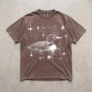 May include: A brown short-sleeve t-shirt featuring a white graphic print of a loon swimming on water. The design includes several white starburst shapes surrounding the bird, creating a celestial effect against the solid brown fabric background.