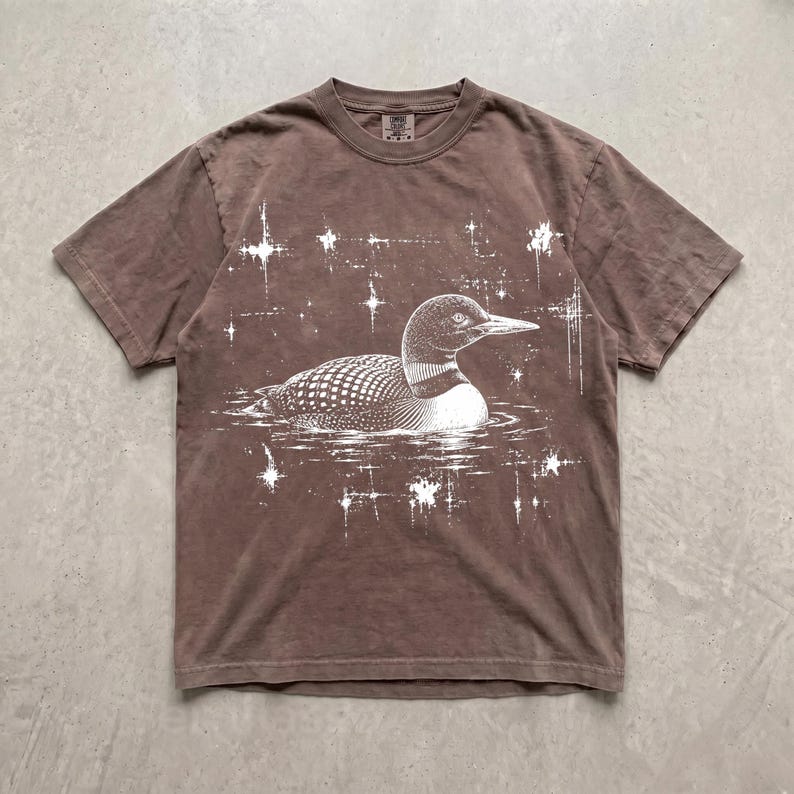 May include: A brown short-sleeve t-shirt featuring a white graphic print of a loon swimming on water. The design includes several white starburst shapes surrounding the bird, creating a celestial effect against the solid brown fabric background.