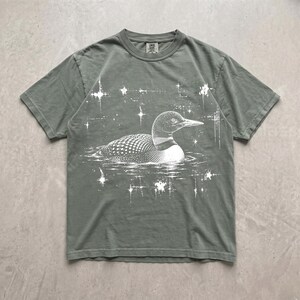 May include: A sage green short-sleeve t-shirt featuring a white graphic print of a loon swimming on water. The design includes stylized starbursts surrounding the bird, creating a celestial effect. The shirt has a crew neckline and a standard fit.