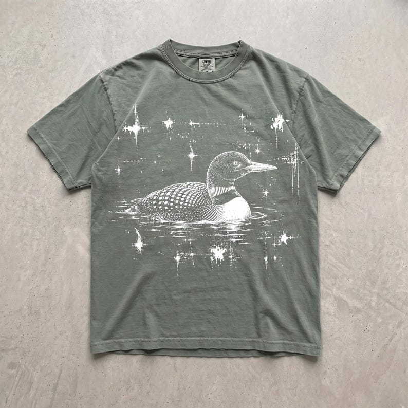 May include: A sage green short-sleeve t-shirt featuring a white graphic print of a loon swimming on water. The design includes stylized starbursts surrounding the bird, creating a celestial effect. The shirt has a crew neckline and a standard fit.