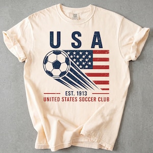 Puede incluir: Una camiseta de manga corta color crema con un gr&aacute;fico de un bal&oacute;n de f&uacute;tbol en movimiento sobre una bandera estadounidense desgastada. El texto dice USA, EST. 1913 y UNITED STATES SOCCER CLUB en azul marino y rojo con aspecto vintage.