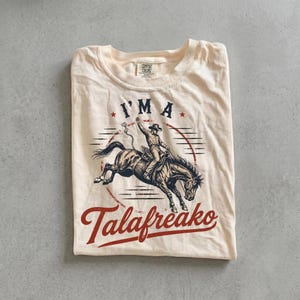 May include: A cream-colored short-sleeve t-shirt featuring a graphic of a cowboy riding a bucking horse. The text above reads I'M A and the text below reads Talafreako in red script. The shirt has a crew neckline and a Comfort Colors brand label.