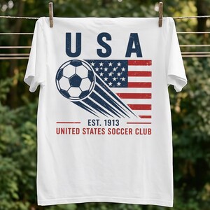 Puede incluir: Una camiseta blanca con un gr&aacute;fico de un bal&oacute;n de f&uacute;tbol en movimiento sobre una bandera estadounidense desgastada. El texto USA aparece en la parte superior, y EST. 1913 UNITED STATES SOCCER CLUB en la parte inferior en letras azul oscuro y rojo.